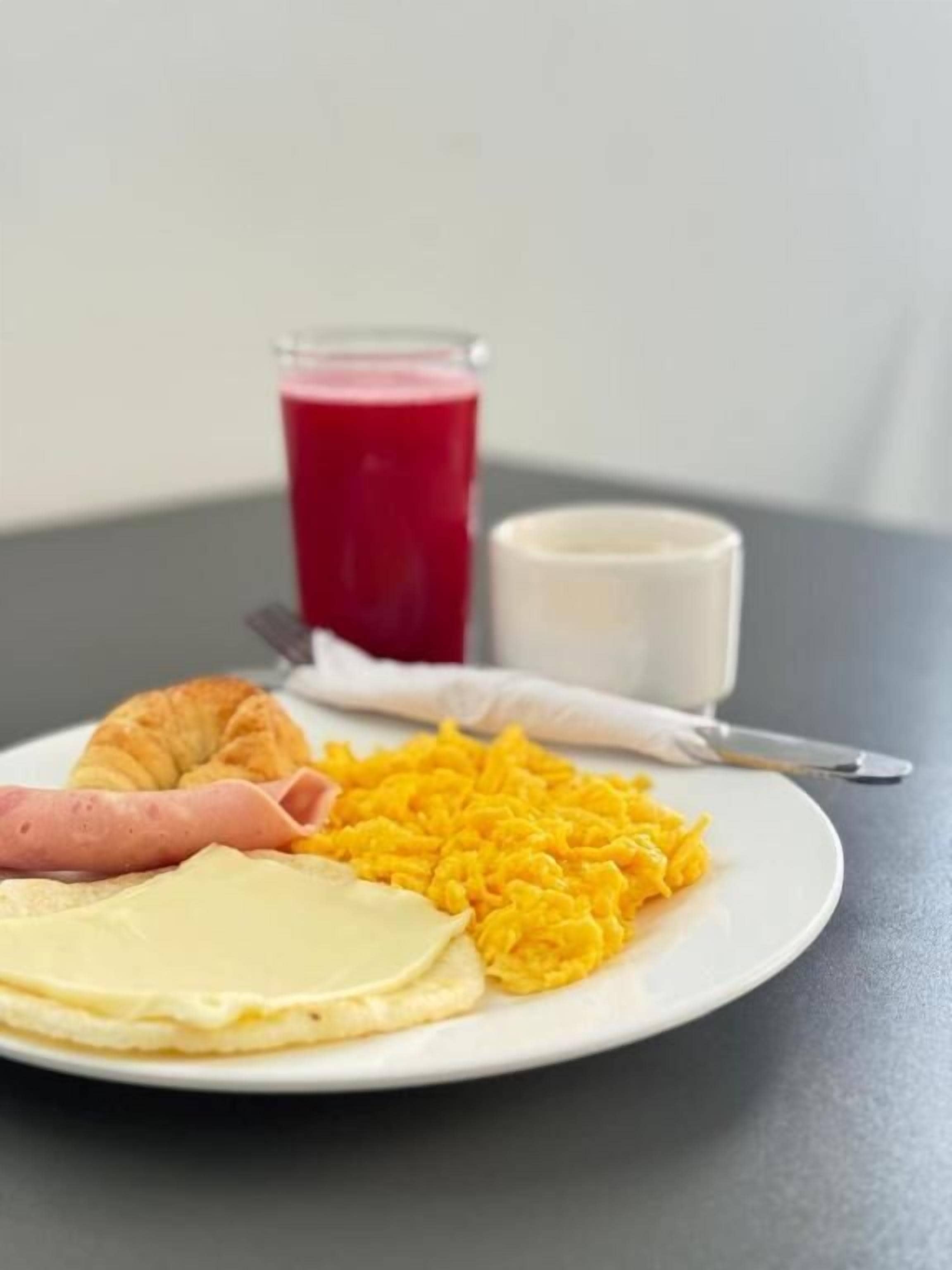 A plate with scrambled eggs, ham, cheese, and a croissant, accompanied by a glass of red juice and a cup of milk on a table.