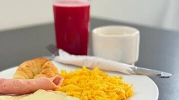 A plate with scrambled eggs, ham, cheese, and a croissant, accompanied by a glass of red juice and a cup of milk on a table.