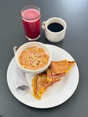 A plate with a sandwich, a bowl of cereal, a glass of juice, and a cup of coffee. - Hotel Gallery By 70 (Medellín)