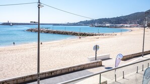 A beach with a clear view of the sea, a rocky breakwater, and a distant cityscape. - Sesimbra Ocean Terrace (Sesimbra)