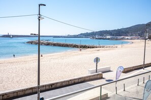 A beach with a clear view of the sea, a rocky breakwater, and a distant cityscape. - Sesimbra Ocean Terrace (Sesimbra)