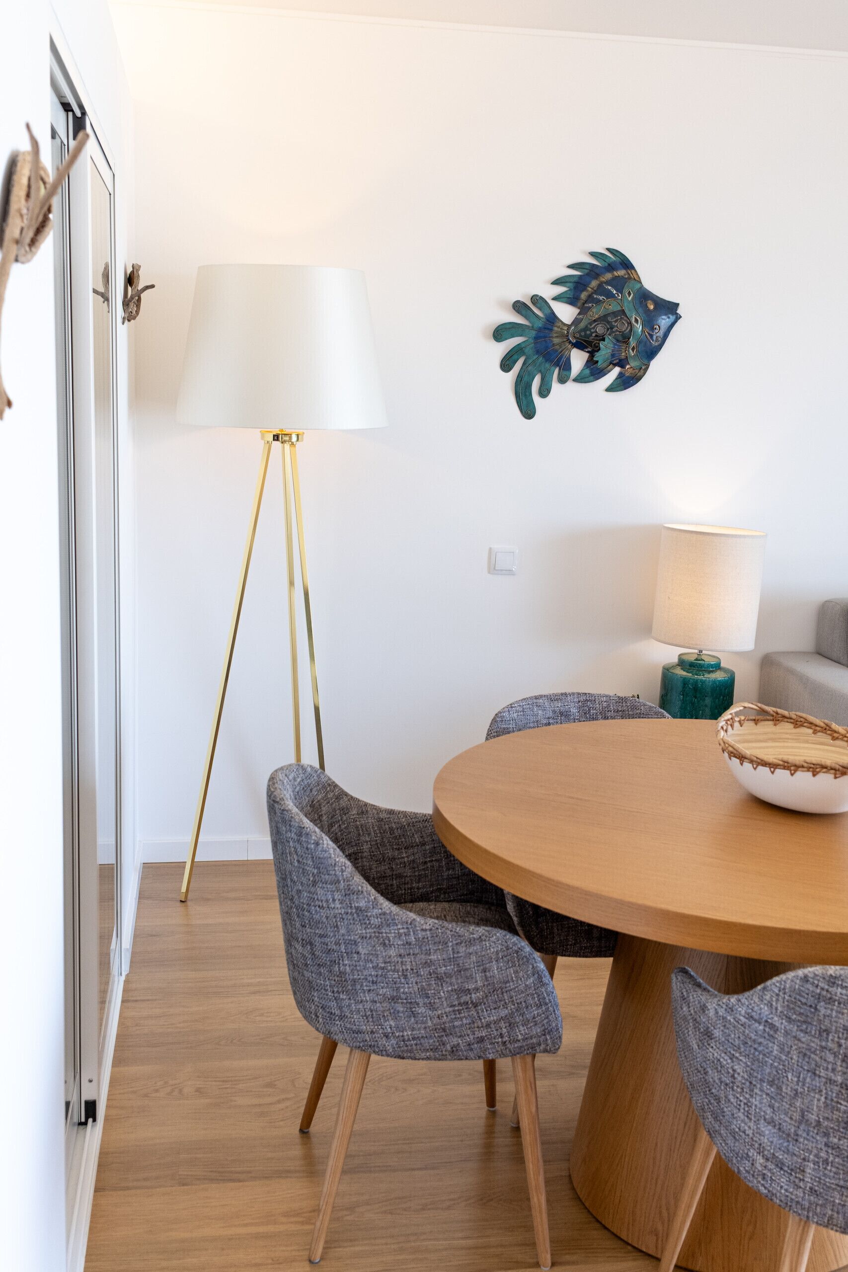 Un coin repas comprenant une table ronde en bois, des chaises à revêtement gris, un lampadaire et une décoration murale représentant des poissons.