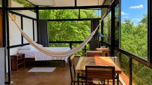 Ein Zimmer mit Bett, Hängematte, Tisch und Stühlen, mit großen Fenstern und Blick auf viel Grün.