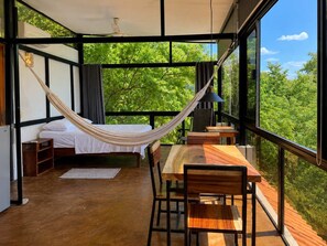 Ein Zimmer mit Bett, Hängematte, Tisch und Stühlen, mit großen Fenstern und Blick auf viel Grün.