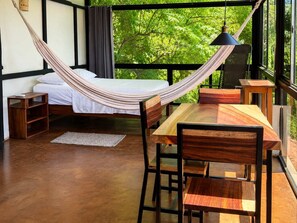 Een hangmatbed met uitzicht op groen, een houten eettafel met stoelen, en een klein nachtkastje.
