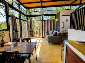 Ein geräumiges Zimmer mit großem Fenster, einem Sofa, einem Esstisch mit Unterlagen und einer Küchenzeile.