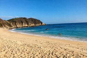 Ein Sandstrand mit klarem, blauem Wasser, felsigen Klippen und zwei Personen im Wasser.
