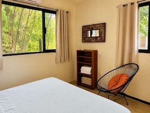 A bedroom with a bed, a chair, a shelf with folded towels, a window with curtains, and a mirror on the wall. - Bungalow doble, cama Queen, cama individual y terraza exterior cocina completa y sofa cama (Cuatunalco)