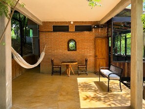 A room with a brick wall, a wooden door, a hammock, two chairs, a table, and a bench.