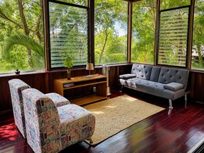 Een kamer met een bank, een gestreept fauteuil, een houten console en een groot raam met zonwering.
