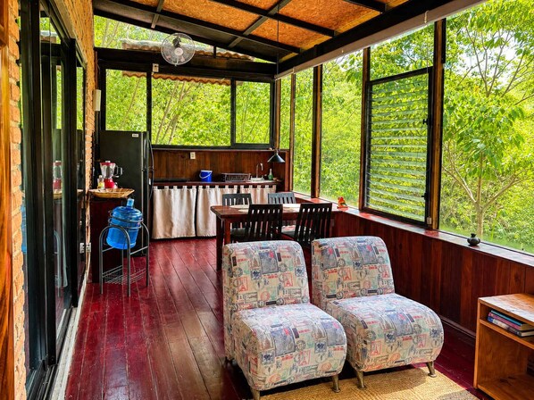 A cozy room with a wooden floor, a dining table, two patterned armchairs, a kitchen area with a sink and a fan, and large windows offering a view of greenery.