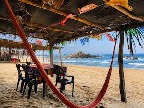 A beachside dining area with a table and chairs under a thatched roof, overlooking the ocean. - Bungalow doble, cama Queen, cama individual y terraza exterior cocina completa y sofa cama (Cuatunalco)