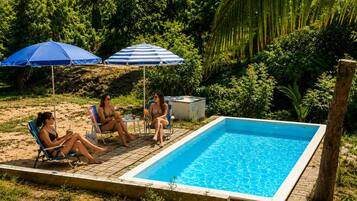 A rectangular swimming pool with a tiled edge, surrounded by a brick patio. There are two blue umbrellas providing shade, and a natural, green backdrop of trees and foliage.