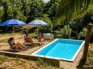 A rectangular swimming pool with a tiled edge, surrounded by a brick patio. There are two blue umbrellas providing shade, and a natural, green backdrop of trees and foliage.