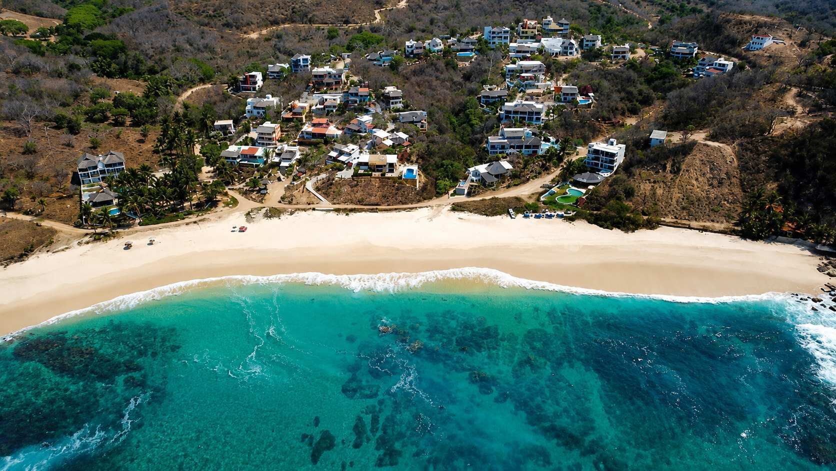 Luftaufnahme eines Küstenresorts mit mehreren Gebäuden, einem Sandstrand und klarem türkisblauem Wasser.