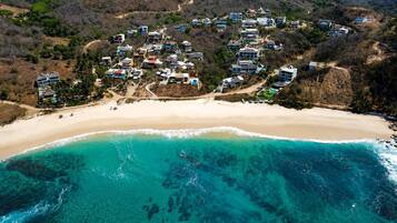 Luftaufnahme eines Küstenresorts mit mehreren Gebäuden, einem Sandstrand und klarem türkisblauem Wasser.