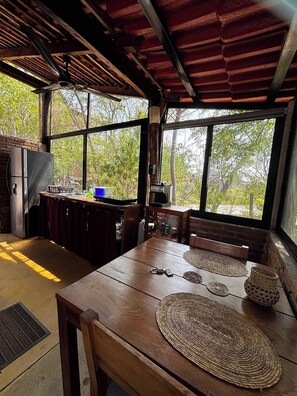 Un coin repas rustique avec une table et des chaises en bois, un ventilateur de plafond et une vue sur les arbres à travers de grandes fenêtres.