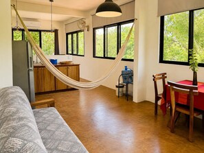 A spacious living area with a hammock, a dining table with chairs, a kitchen area, and large windows with a view of greenery. - Valeria Bungalow doble, cama Queen, cocina completa y sofa cama (Cuatunalco)