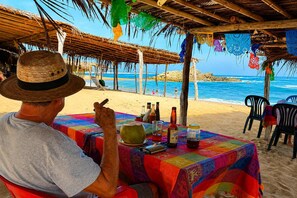 Eine Strandansicht mit einem für zwei Personen gedeckten Tisch, mit einer bunten Tischdecke und verschiedenen Flaschen.