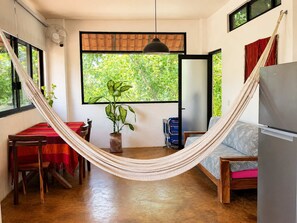 A hammock suspended between two windows, a dining table with chairs, a sofa, a refrigerator, and a potted plant. - Valeria Bungalow doble, cama Queen, cocina completa y sofa cama (Cuatunalco)