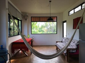 Ein Zimmer mit einer Hängematte, einem Tisch, einer Couch, einem großen Fenster mit Blick auf Bäume und einem Deckenventilator.