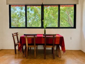 A dining area with a table set for two, a window with a view of trees, and a vase with a plant on the table. - Valeria Bungalow doble, cama Queen, cocina completa y sofa cama (Cuatunalco)