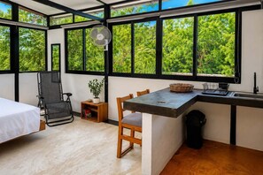 A compact room with a bed, a desk, a chair, a small table, a fan, and a window with a view of greenery. - Bungalow doble, cama Queen, cocina completa. (Cuatunalco)