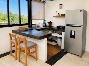 A compact kitchen with a refrigerator, a sink, and a window overlooking greenery. - Bungalow doble, cama Queen, cocina completa. (Cuatunalco)