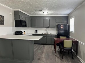 A kitchen with grey cabinets, a black refrigerator, a coffee maker, and a dining table with chairs.
