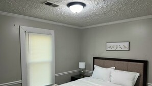 A bedroom with a bed, a nightstand, a lamp, a window with blinds, and a ceiling light fixture.