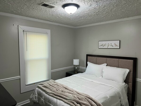 A bedroom with a bed, a nightstand, a lamp, a window with blinds, and a ceiling light fixture.