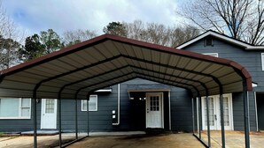 A covered garage with a metal roof and a concrete driveway leading to a house with a white door.