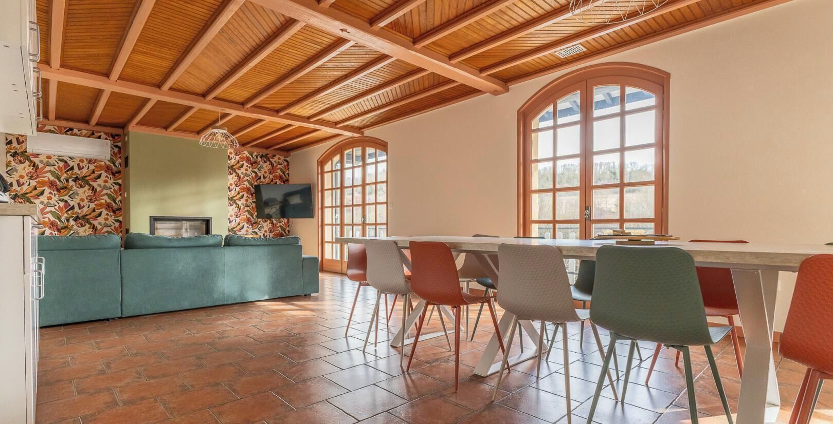 A spacious room with a wooden ceiling, a dining table with chairs, a sofa, and a large window.