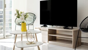 Un salon moderne avec un téléviseur à écran plat, une table ronde blanche ornée d’un vase jaune et deux chaises noires en fil de métal.