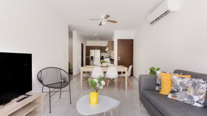 Un salon moderne doté d’un téléviseur à écran plat, d’un coin repas avec une table et des chaises, d’un canapé gris agrémenté de coussins décoratifs et d’un ventilateur de plafond.