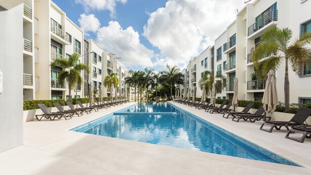 Une piscine entourée de chaises longues et de palmiers, le tout bordé d’immeubles modernes.
