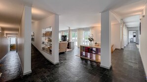 Un hall d’entrée spacieux, doté d’un sol en pierre sombre, de murs blancs et d’un espace salon central avec un téléviseur et un vase.