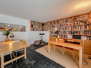 Une pièce avec une table en bois, des chaises, une bibliothèque remplie de livres, un bureau avec une lampe et une petite plante décorative.