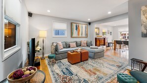 A spacious living room with a grey sofa, ottomans, a patterned rug, and a television. - Spacious 3BR/2.5BA · Fenced Backyard Oasis · Pets OK (Washington)