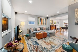 Ein geräumiges Wohnzimmer mit grauem Sofa, Poufs, einem gemusterten Teppich und einem Fernseher.
