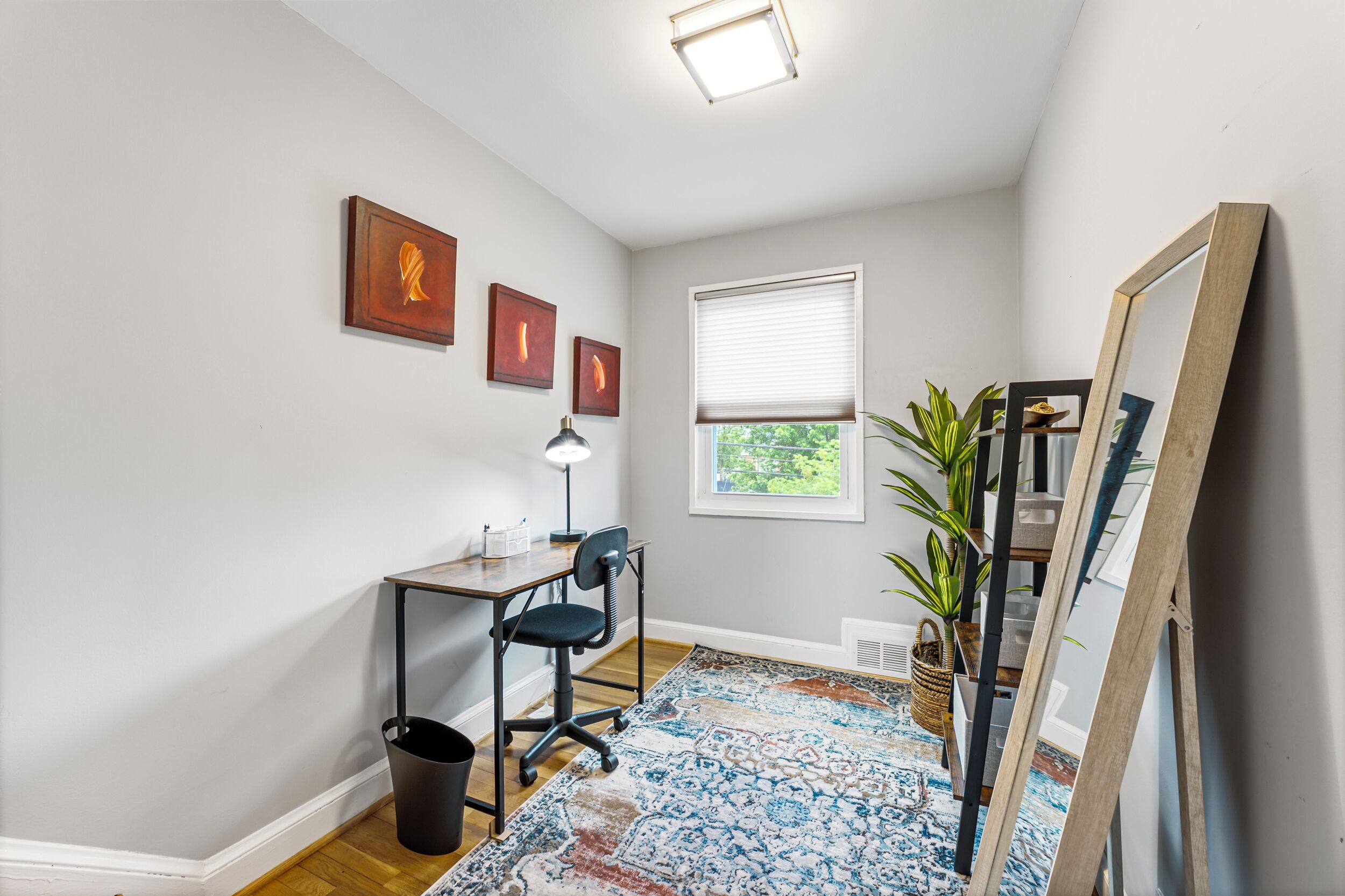 Una habitación con escritorio, silla, lámpara, ventana y una obra de arte en la pared.