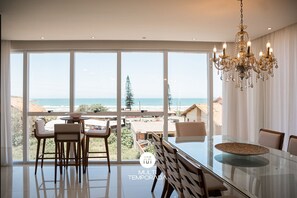 A modern dining area with a glass table, chairs, and a chandelier. - Apartamento Simon Vista Mar - Imbituba (Imbituba)