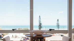 Een kamer met een groot raam met uitzicht op een strand, een ronde houten tafel met een kom, en twee stoelen.