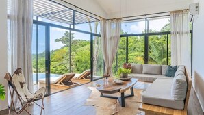 A modern living room with a large window, a sofa, a coffee table, and a chair. - Brand-New Jungle Hideaway with Ocean View (Sámara)