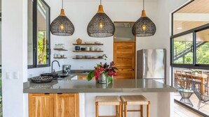 A modern kitchen with a central island, pendant lights, and a view to an outdoor area. - Brand-New Jungle Hideaway with Ocean View (Sámara)
