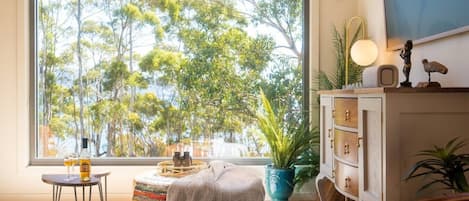 Um quarto com uma janela grande, uma mesa redonda pequena, um banco, um armário e uma planta.