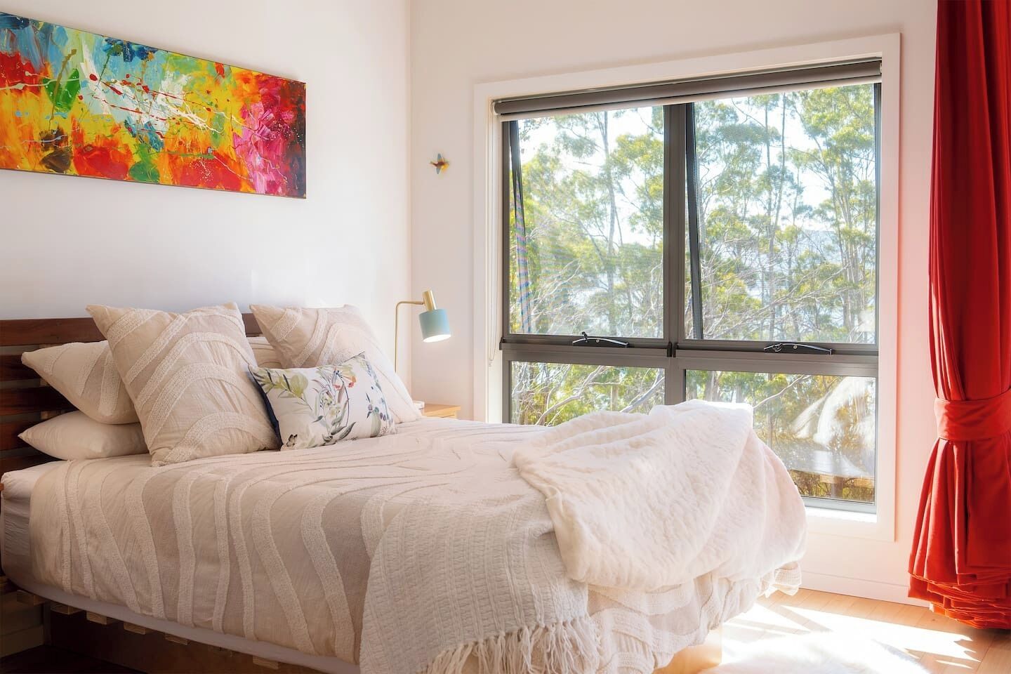 Une chambre avec une grande fenêtre, un lit recouvert de plusieurs oreillers, un tableau abstrait aux couleurs vives accroché au mur et un rideau rouge.