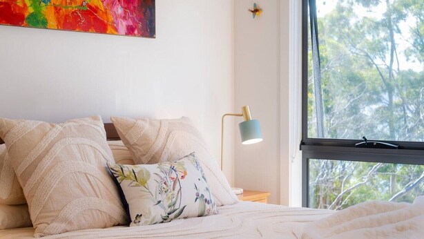 Un lit avec un coussin à motifs floraux, une lampe et une fenêtre donnant sur des arbres.