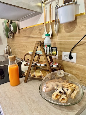 A kitchen counter with a glass display case of pastries, a bottle of orange juice, and a wooden bread rack with bread. - Domus Auri Vatican Jacuzzi Boutique Stay (Roma)