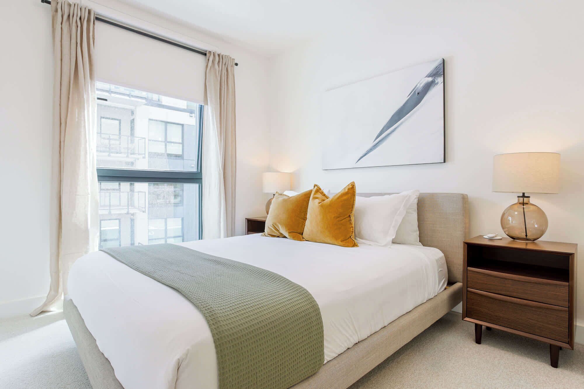 A hotel room with a large bed, two bedside tables, a window with curtains, a wall-mounted lamp, and a painting.